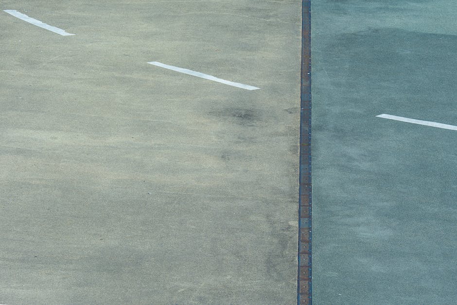 An aerial view of a large concrete parking lot divided into two sections by a central metal drain cover. The left section shows darker, weathered concrete with visible dirt and tire marks, while the right section appears cleaner with a brighter, more uniform surface. White painted lines designate parking spaces, with one line partially visible in the upper left corner. The lighting suggests daytime, with natural light enhancing the contrast between the two areas. This image emphasizes surface cleaning and maintenance aspects related to commercial parking areas, reflecting processes of deep cleaning and surface hygiene, aligning with the services of Carpet Cleaners Holland Park, a specialist in surface cleaning and sanitisation for both residential and commercial clients.