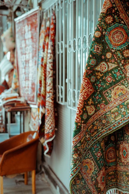 Indoor display of intricately patterned oriental rugs hanging on white metal grid panels and draped over a wooden chair, showcasing vibrant colors including red, green, orange, and beige. The rugs feature detailed floral and geometric designs on woven fabric surfaces. The scene is lit by natural light, highlighting the textures and details of the rugs. In the background, blurred elements of a retail or showroom environment are visible, with additional rugs and furniture. This setting illustrates surface cleaning and maintenance processes typical of professional rug care services offered by Carpet Cleaners Holland Park, specializing in deep cleaning and sanitisation of domestic and commercial rugs in the Holland Park area.