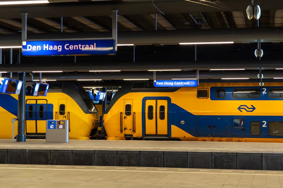 Interior view of Den Haag Centraal railway station showing two modern, double-decker trains painted in yellow and blue parked at the platform. The platform is clean and well-lit, with a smooth concrete surface and minimal debris. Overhead, illuminated signage displays the station name, 'Den Haag Centraal,' in white text on a blue background, suspended from the ceiling. The station's ceiling features a structured, grid-like pattern with horizontal lighting fixtures providing even illumination across the area. In the background, there are additional digital screens and small seating areas, emphasizing a tidy and orderly environment typical of professional public transportation hubs. The sleek design and pristine condition of the trains and platform reflect regular cleaning and maintenance, aligning with high standards of hygiene associated with professional cleaning services like those offered by Carpet Cleaners Holland Park.