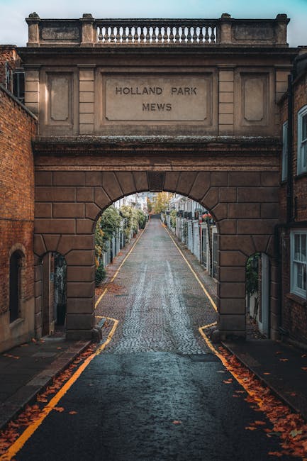 The image depicts the entrance to Holland Park Mews, featuring a large stone archway with the inscription 'HOLLAND PARK MEWS' engraved on it. The arch is constructed of brownish stone bricks with a decorative balustrade along the top. The narrow street beyond the arch is paved with cobblestones and lined with residential buildings and lush greenery. The scene is illuminated by natural daylight, with fallen leaves scattered along the edges of the street, creating a calm and picturesque atmosphere. This setting exemplifies a typical quiet, well-maintained lane in Holland Park, showcasing classic architectural features and tranquil urban environment. The image is relevant to the cleaning service offered by Carpet Cleaners Holland Park, emphasizing community and local charm, alongside the importance of maintaining clean and hygienic residential and commercial spaces.