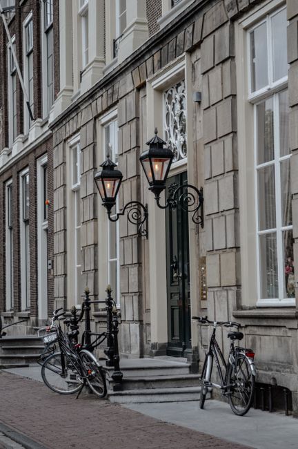 Exterior view of a historic building with a stone facade featuring decorative architectural details. Two vintage-style black lanterns with glass enclosures are mounted on ornate iron brackets beside the dark green front door, which has a decorative iron grille and a small window above. Three bicycles are parked on the sidewalk in front of the building, leaning against the iron railing. The area is well-lit by natural daylight, highlighting the clean and well-maintained appearance of the building's exterior. This scene exemplifies the charming, classic urban architecture common in Holland Park, demonstrating attention to exterior maintenance that complements professional cleaning services offered by Carpet Cleaners Holland Park for residential and commercial properties.