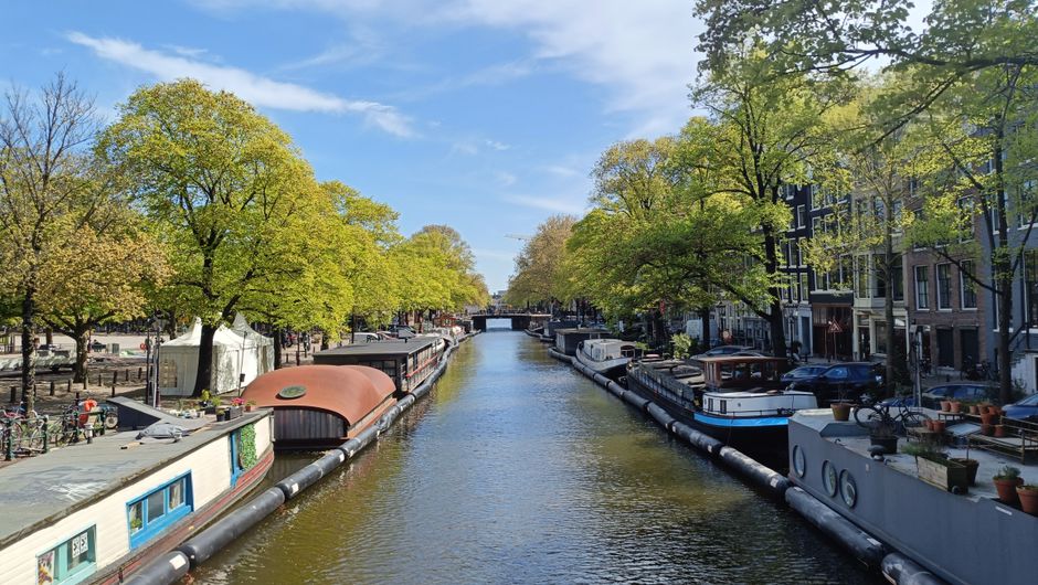 A scenic view of a canal in Holland Park with several narrowboats moored along both sides, surrounded by green-leaved trees under a bright blue sky with scattered clouds. Residential buildings with large windows and balconies line the canal, and some potted plants are visible on the balconies. The water in the canal appears calm and reflective, creating a peaceful atmosphere. The image captures an outdoor setting typical of the Holland Park terraces, emphasizing the natural surroundings and urban waterways, indicative of a clean, well-maintained environment ideal for surface cleaning and maintenance by companies like Carpet Cleaners Holland Park.