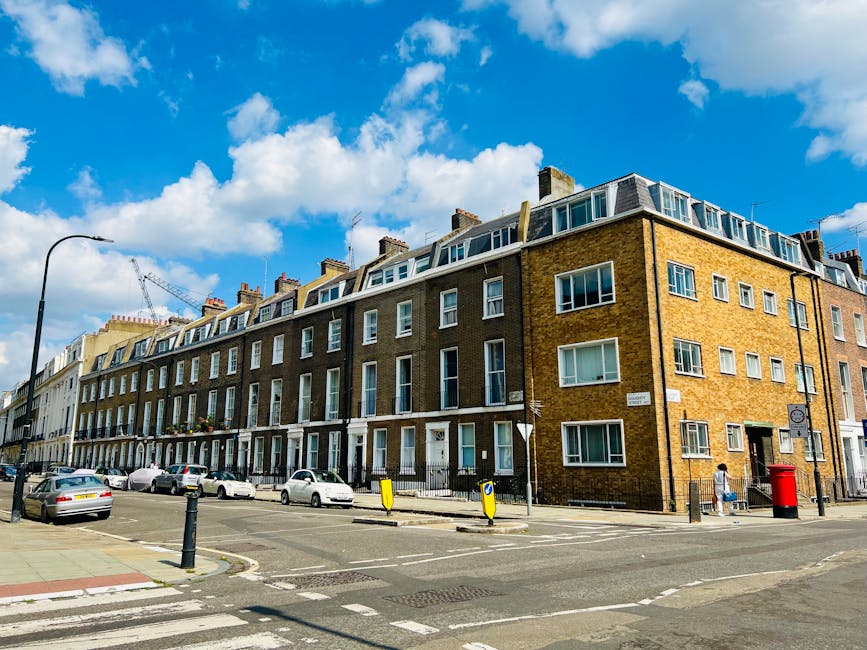 Photograph of a multi-story residential building with a brick facade, large windows, and modern dormer windows on the top floor, situated on a busy city street under a bright blue sky with scattered w