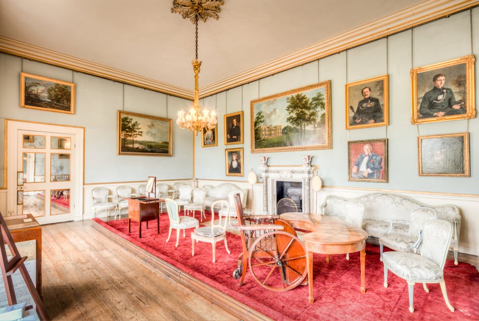 A spacious, well-lit room with light blue walls decorated with multiple framed landscape and portrait paintings arranged around a central white fireplace with ornate detailing. The room features antique-style white and pale green upholstered chairs and a matching sofa, arranged around a polished wooden table. A deep red area rug covers the wooden floor, and a brass chandelier hangs from the ceiling, providing elegant lighting. The room appears clean and maintained, with polished surfaces and neatly arranged furnishings, indicative of professional domestic cleaning or surface cleaning by Carpet Cleaners Holland Park, as seen on the webpage for Melbury Road upholstery cleaning for period houses.