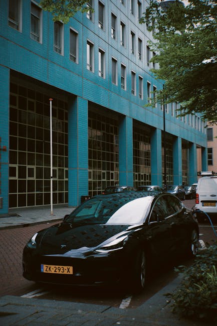 A black electric car parked on a street in front of a modern blue office building with large glass windows. The vehicle is sleek with a glossy finish, reflecting the surrounding environment, and is si